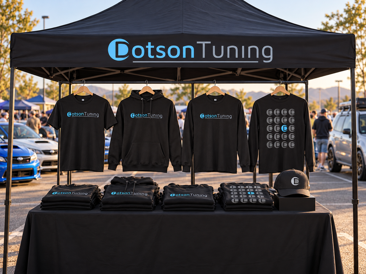 A black Dotson Tuning merchandise canopy at a Subaru car meet, with four shirts and hoodies hanging above a table and a baseball cap on display.